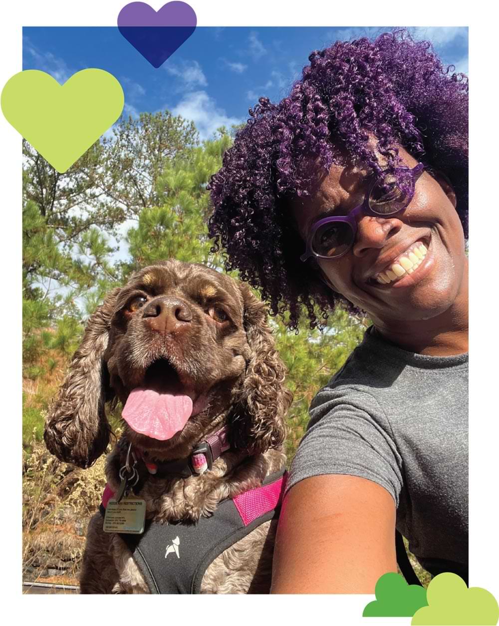 Orazie Cook smiles brightly while taking and outdoor selfie with a dark brown American Cocker Spaniel that wears a pink and black harness