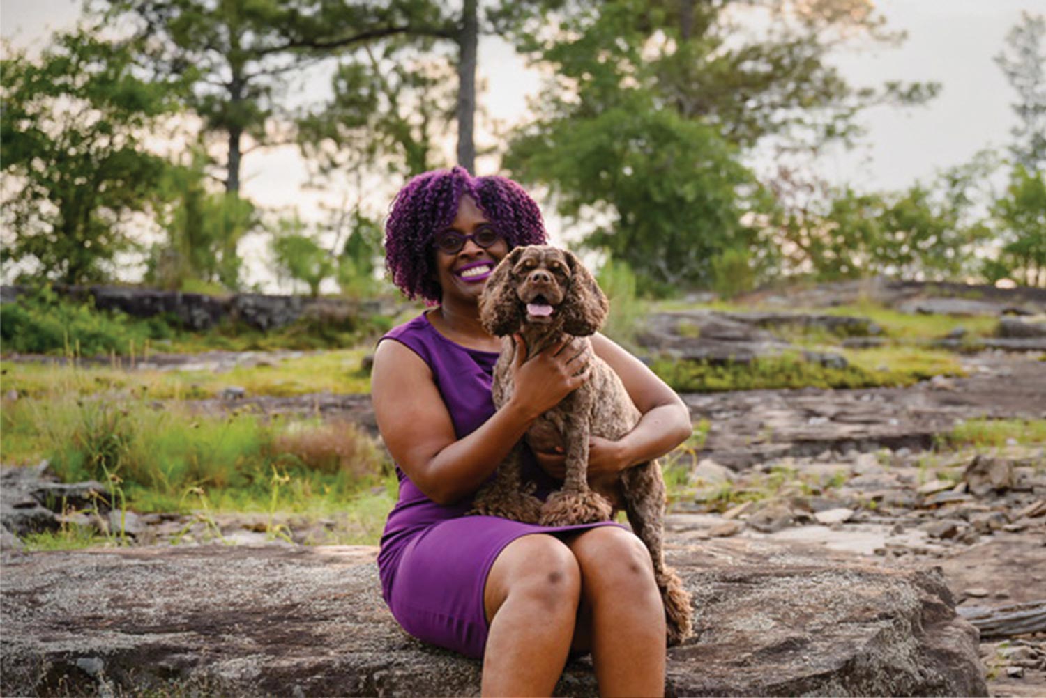 Orazie Cook, wearing a purple dress that matches her vibrant purple hair and glasses, smiles while sitting on a wide low boulder with a dark brown American Cocker Spaniel leaning on her lap