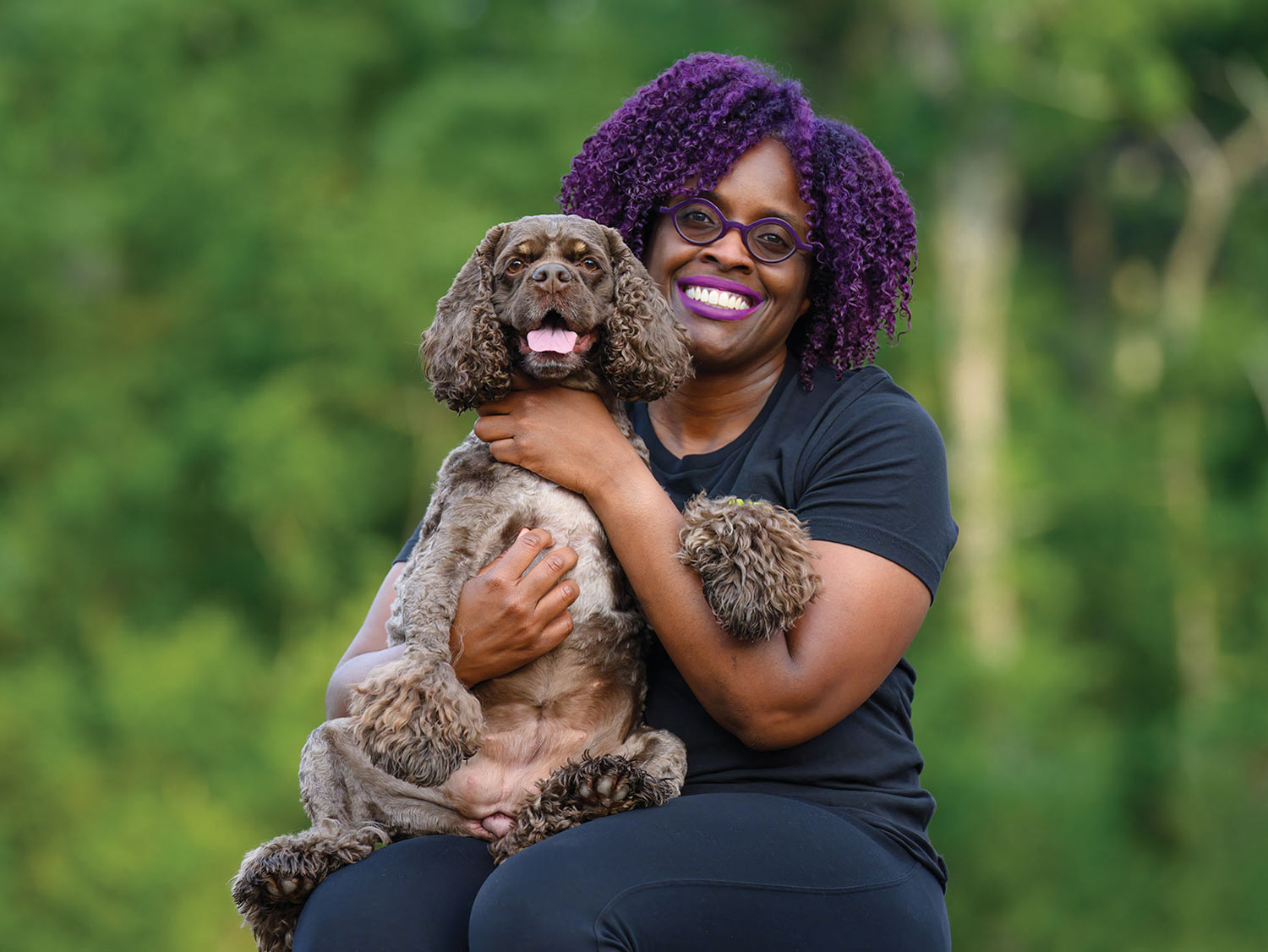 Orazie Cook with her purple hair, lipstick and round thick rimmed glasses is photographed smiling while holding a dark brown American Cocker Spaniel on her lap