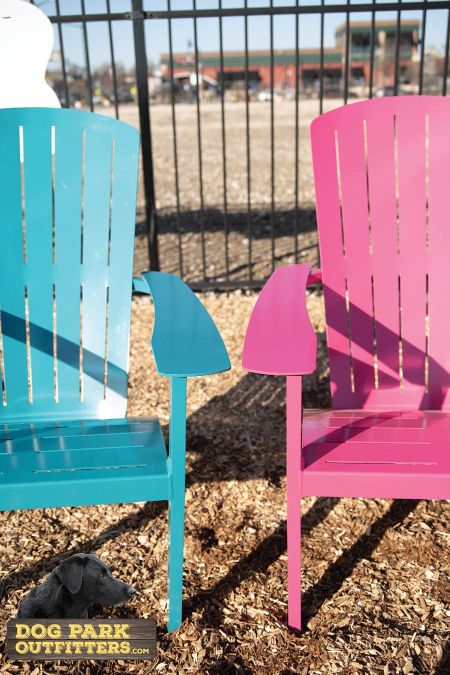Close-up portrait photograph view of The Dog Park Lounge Chairs side by side (blue on the left, pink on the right) from Dog Park Outfitters as the chairs are outside somewhere on top of dark/light brown wood chips surface nearby a black fence