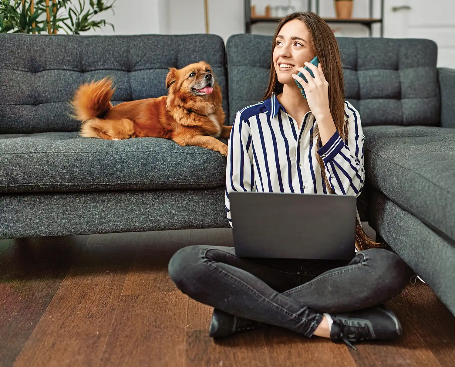 woman sitting cross-legged on the floor and talking on the phone with a laptop on her lap and a dog on the couch beside her