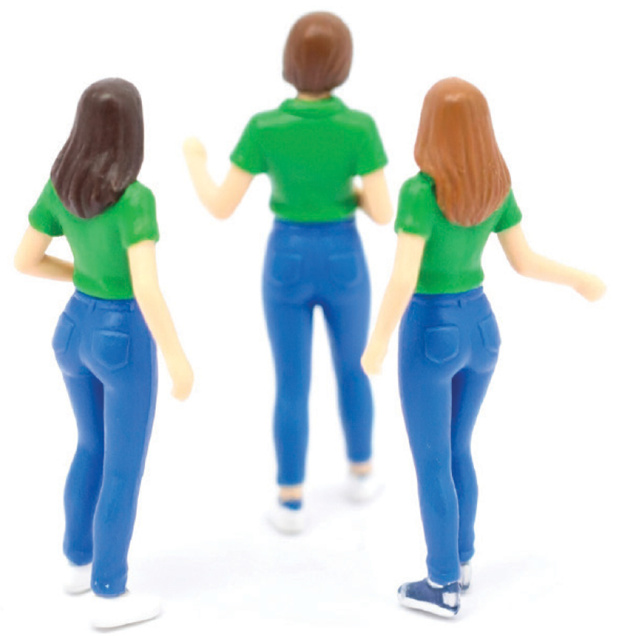 Three miniature figurines of women, viewed from the back, stand wearing green shirts and blue pants.