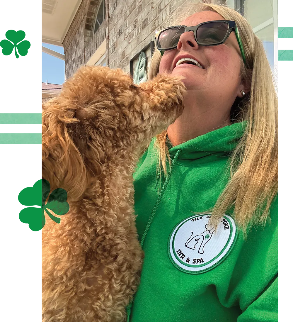 A woman in a green "Woof Tone Inn & Spa" hoodie smiling as a curly-haired dog nuzzles her chin.