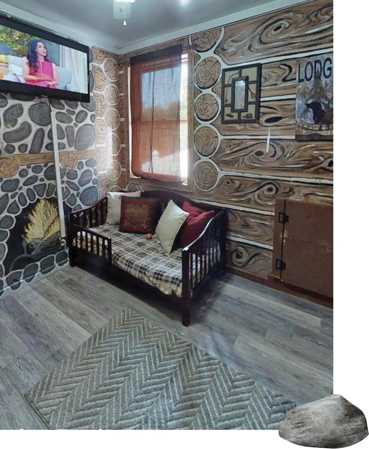 Cozy cabin-themed pet suite featuring log-patterned walls, a small daybed with throw pillows, a wall-mounted television, and a chevron area rug.
