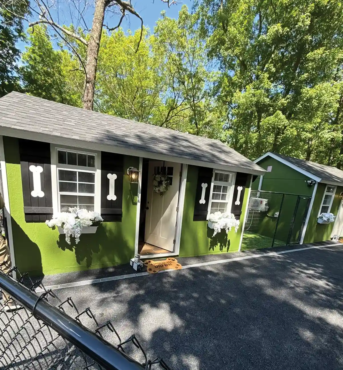 Green cottage-style dog suite featuring window flower boxes, dark shutters with white bone accents, and a bone-shaped doormat at the open door.
