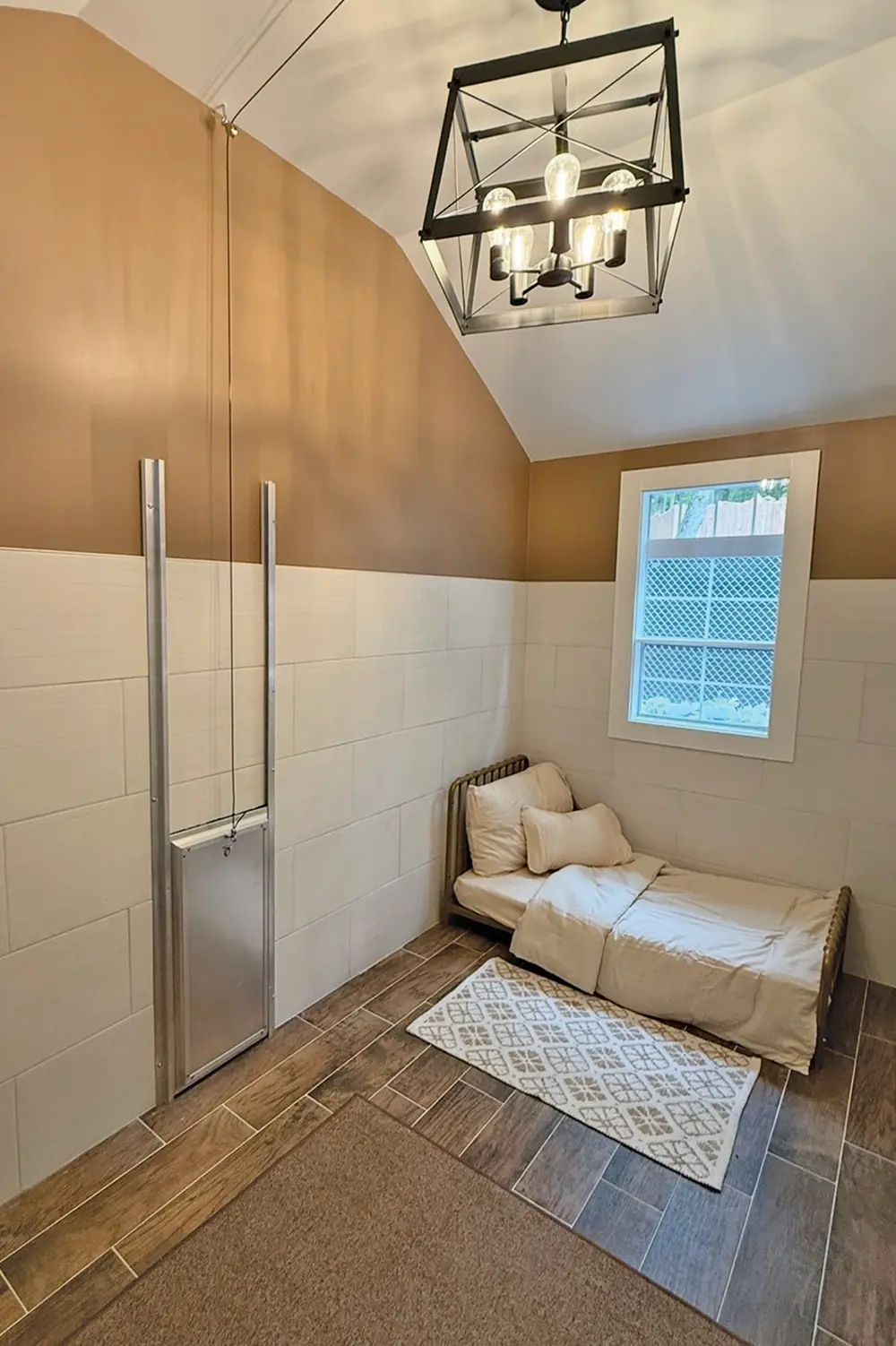 Interior of a luxury dog boarding suite featuring a small bed with pillows, a sliding dog door, tile flooring, a patterned rug, and a modern lantern light fixture.